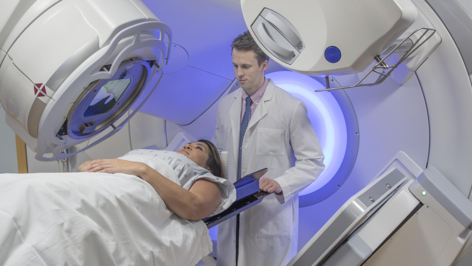 A lady is lying under a radiotherapy machine. A doctor is standing behind her head