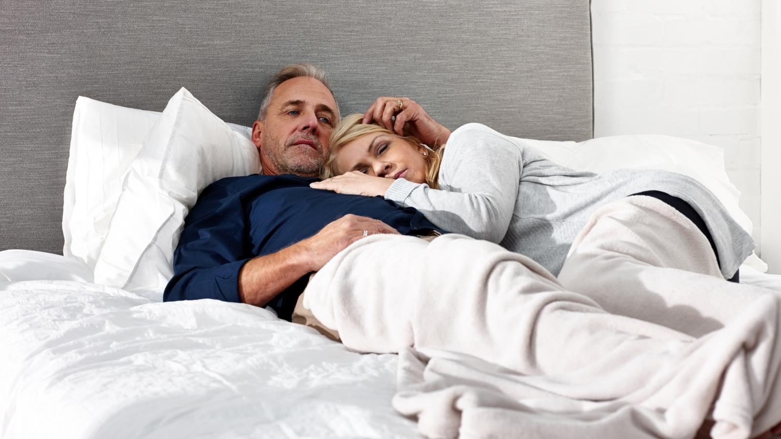 Mature man and woman cuddling in bed. The woman looks a bit worried.