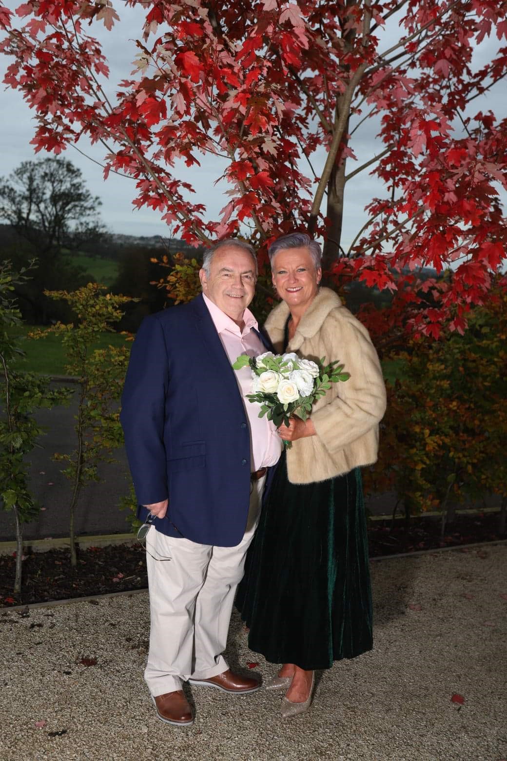 Maggie Donnelly pictured with her husband Pat in front of a tree with red leaves. Maggie is holding a bouquet of white flowers.