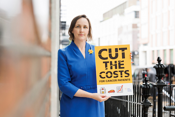 Woman in blue holding yellow placard reading 'cut the costs for cancer patients'