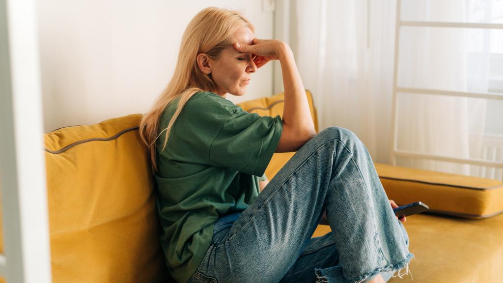 A woman in her thirties sits on a sofa holding her head looking tired or unhappy