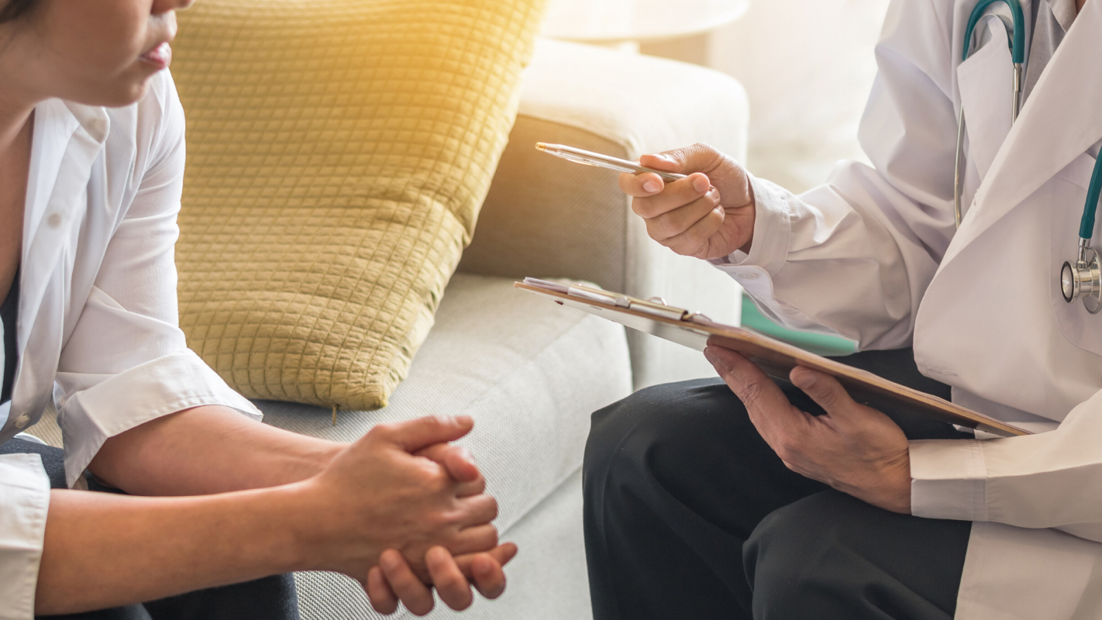 A woman on a sofa is sitting with her hands clasped. A doctor with a clipboard is talking to her.