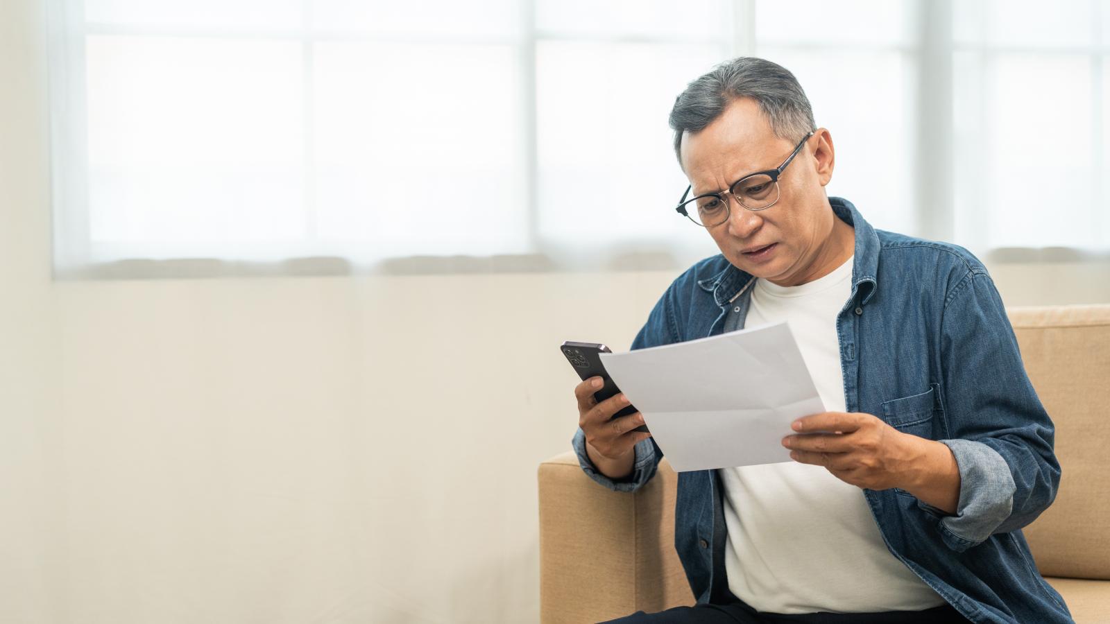 An Asian man in his fifties on a sofa. He is looking concerned holding a piece of paper and a phone